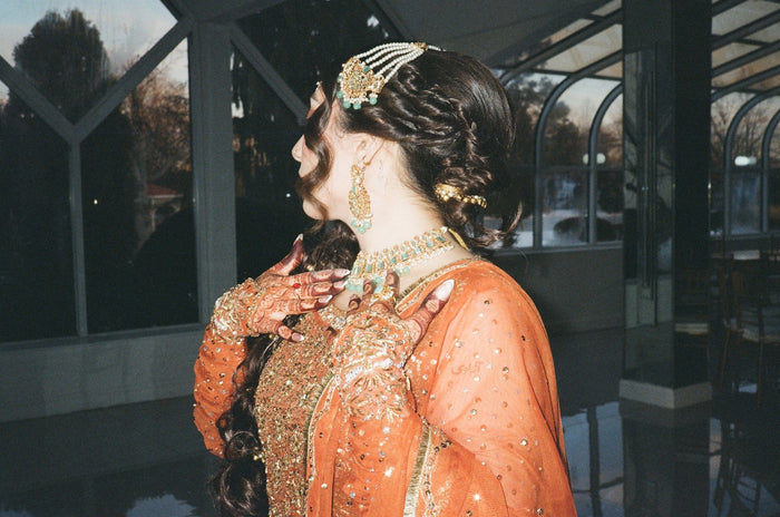 A wedding guest in a rust-orange outfit wearing a green-stone matha patti, a statement chandbali earring and an emerald-and-gold choker, with mehndi-decorated hands